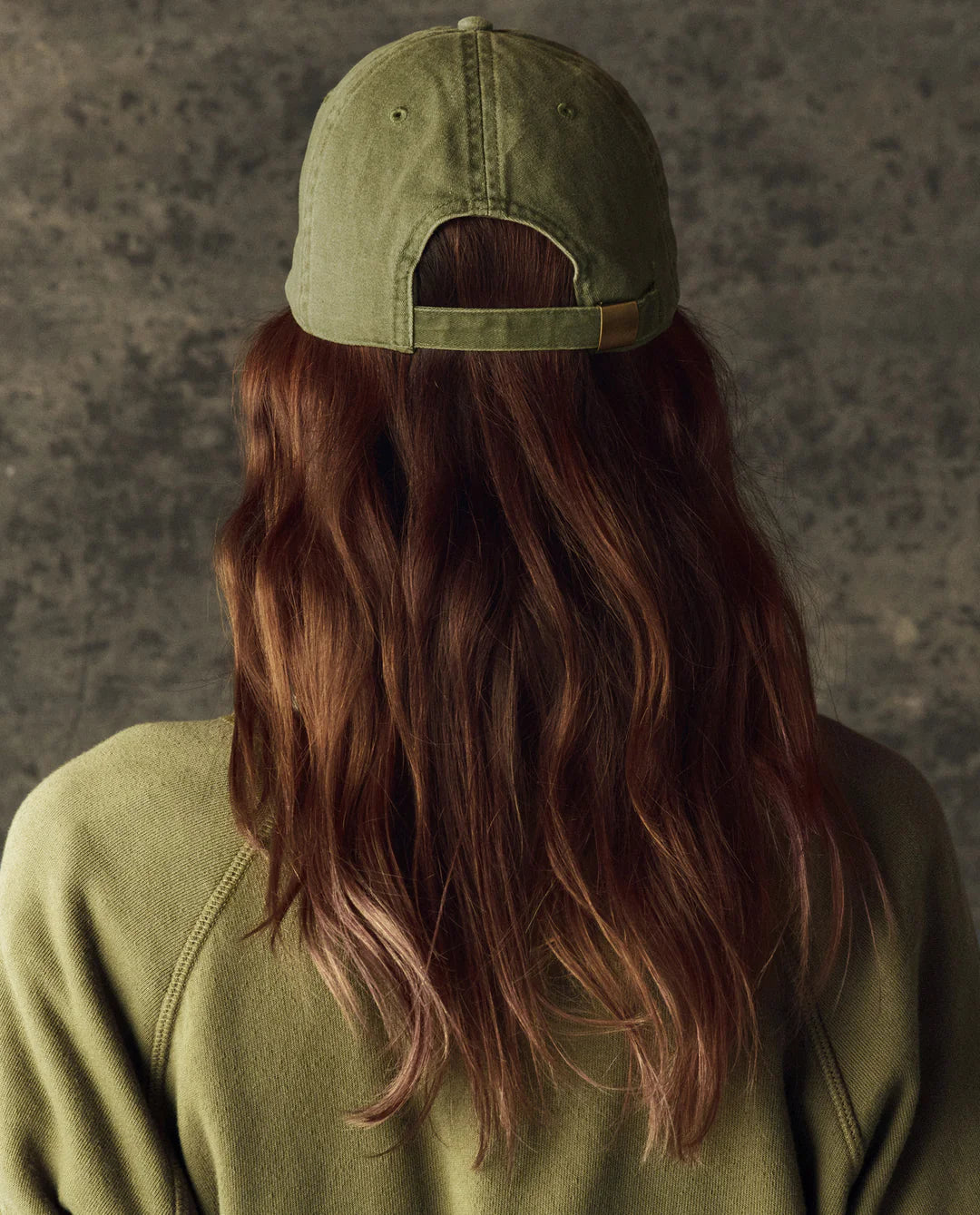 A person with long, wavy brown hair wears The Great Inc.'s Classic Baseball Hat and a matching olive green sweater, facing away from the camera against a textured gray background.