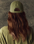 A person with long, wavy brown hair wears The Great Inc.'s Classic Baseball Hat and a matching olive green sweater, facing away from the camera against a textured gray background.