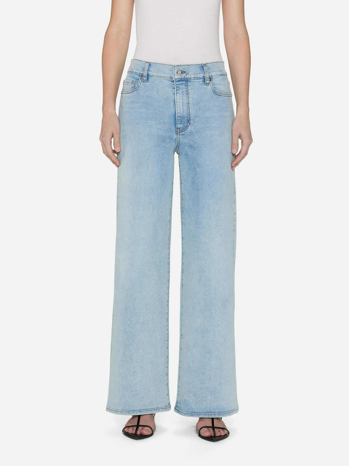 A person is wearing FRAME's Le Slim Palazzo light blue wide-leg jeans, crafted from sustainable cotton in a chic palazzo silhouette with a high rise and two front pockets. They wear a white sleeveless top and black open-toe sandals against a white background.