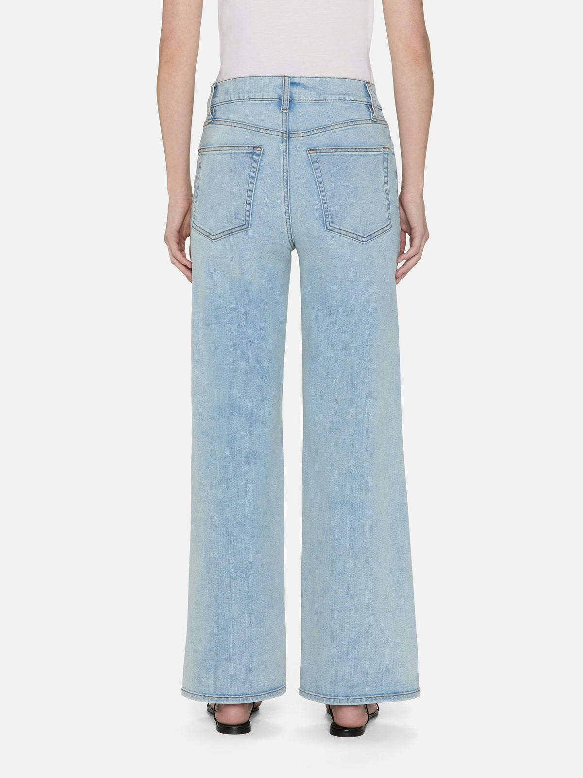 A person stands against a white background in FRAME's Le Slim Palazzo jeans, featuring a high-rise fit, light blue hue, sustainable cotton blend, wide-leg palazzo silhouette, and two back pockets. They pair the jeans with black heels for an effortlessly chic look.