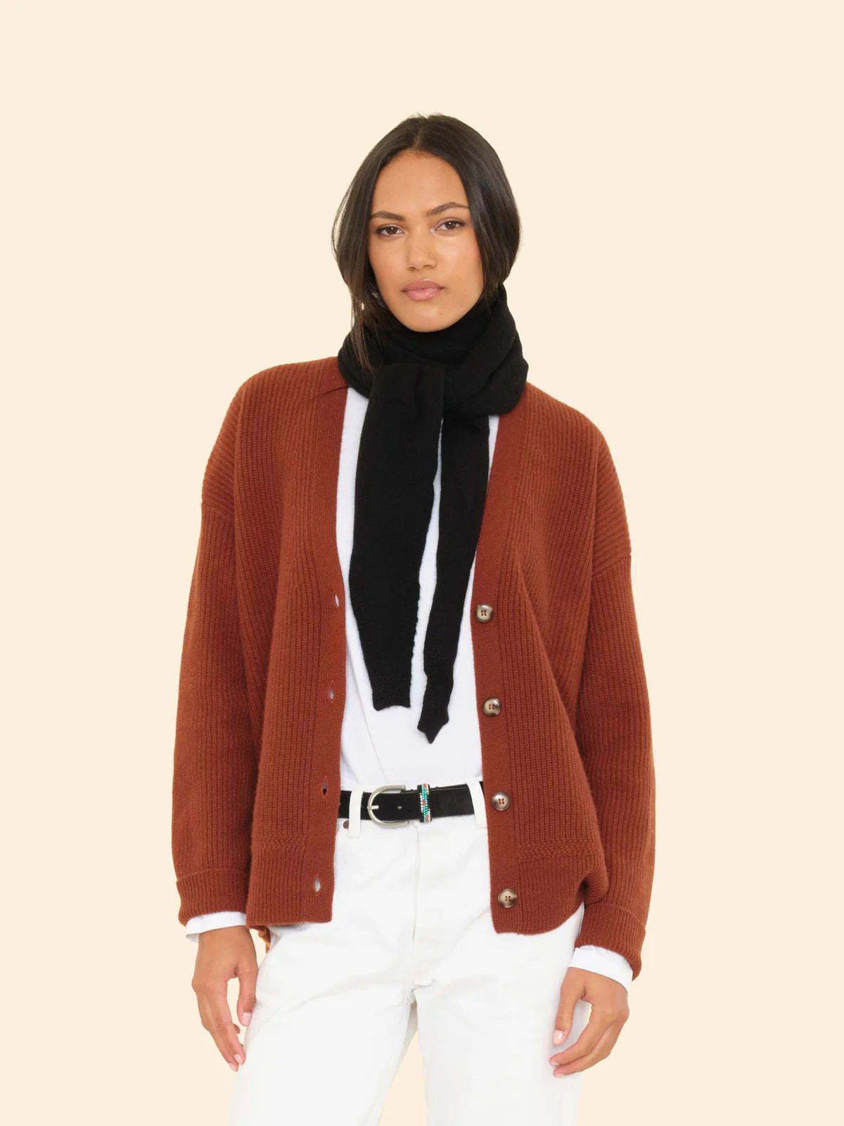 A woman with straight dark hair and a relaxed expression stands against a light background, wearing a brown cardigan, white shirt and pants, black belt, and the ultra-soft Le Petit Scarf draped around her neck.