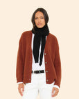 A woman with straight dark hair and a relaxed expression stands against a light background, wearing a brown cardigan, white shirt and pants, black belt, and the ultra-soft Le Petit Scarf draped around her neck.