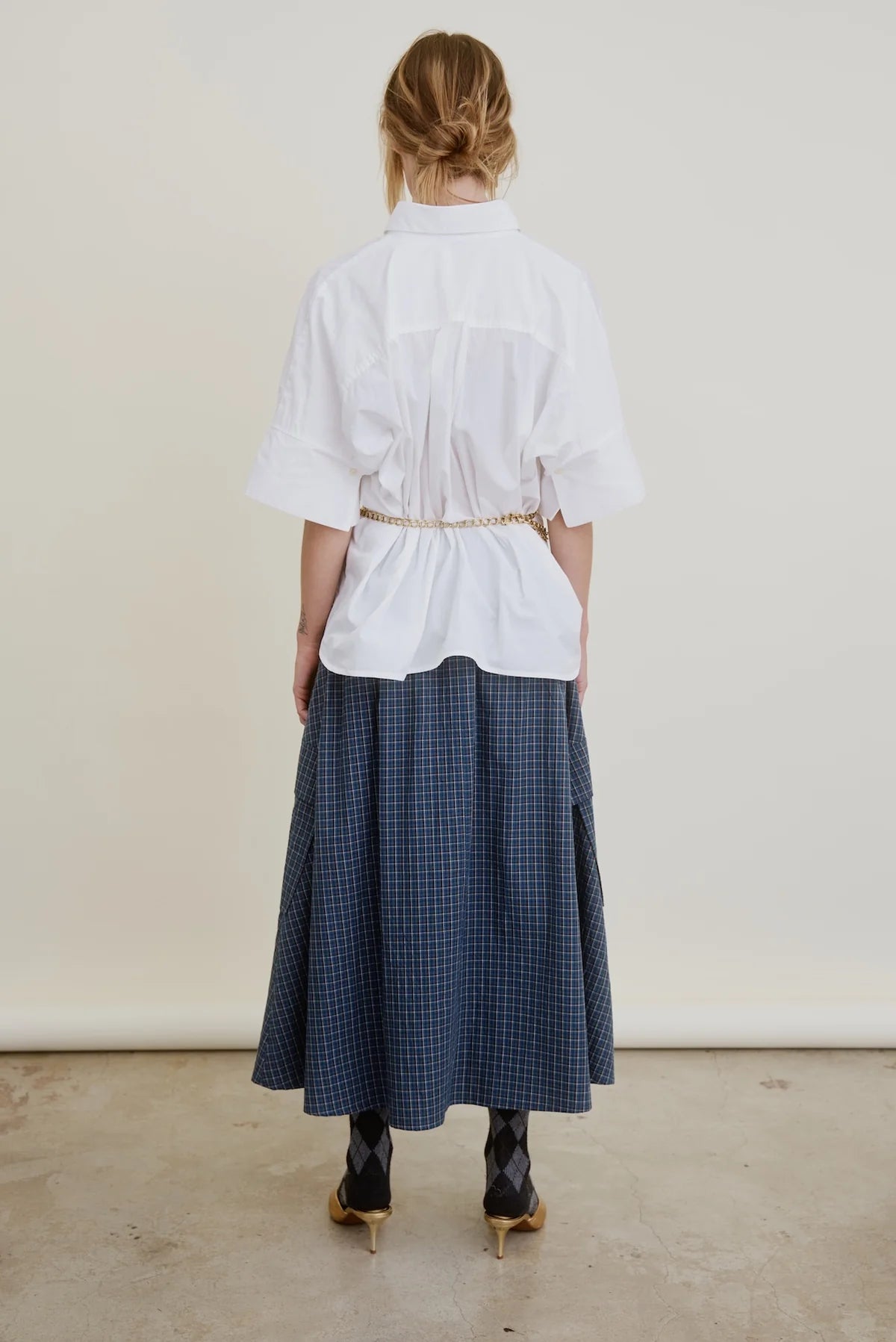 A person stands facing away, wearing the Aquarius Cocktail Le Cargo Skirt in blue plaid, a loose white Japanese cotton poplin shirt cinched with a belt, black argyle socks, and heels. Their hair is tied back against a neutral background.