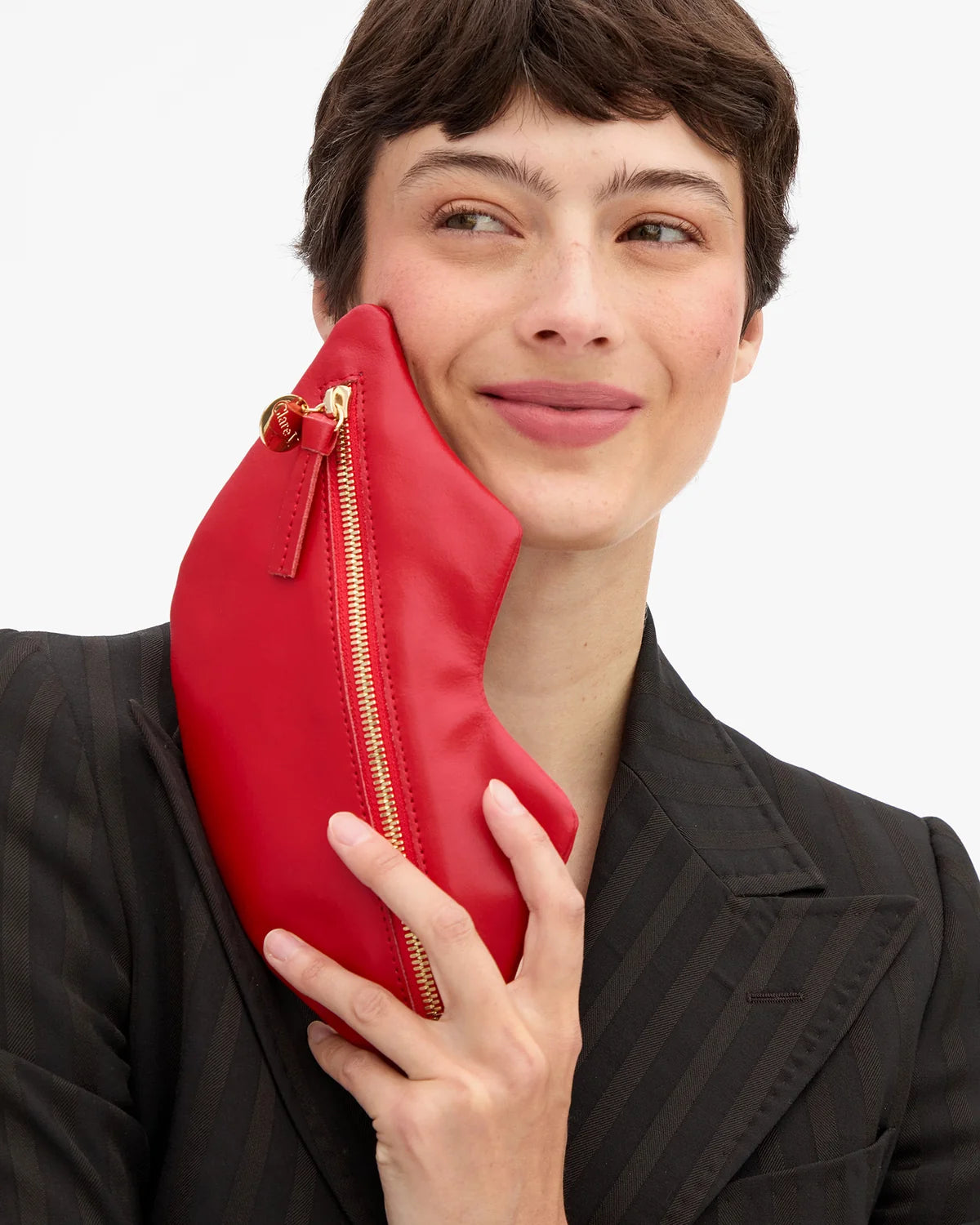 Clare Vivier Lips Flat Clutch in red held against a cheek, featuring a gold zipper.