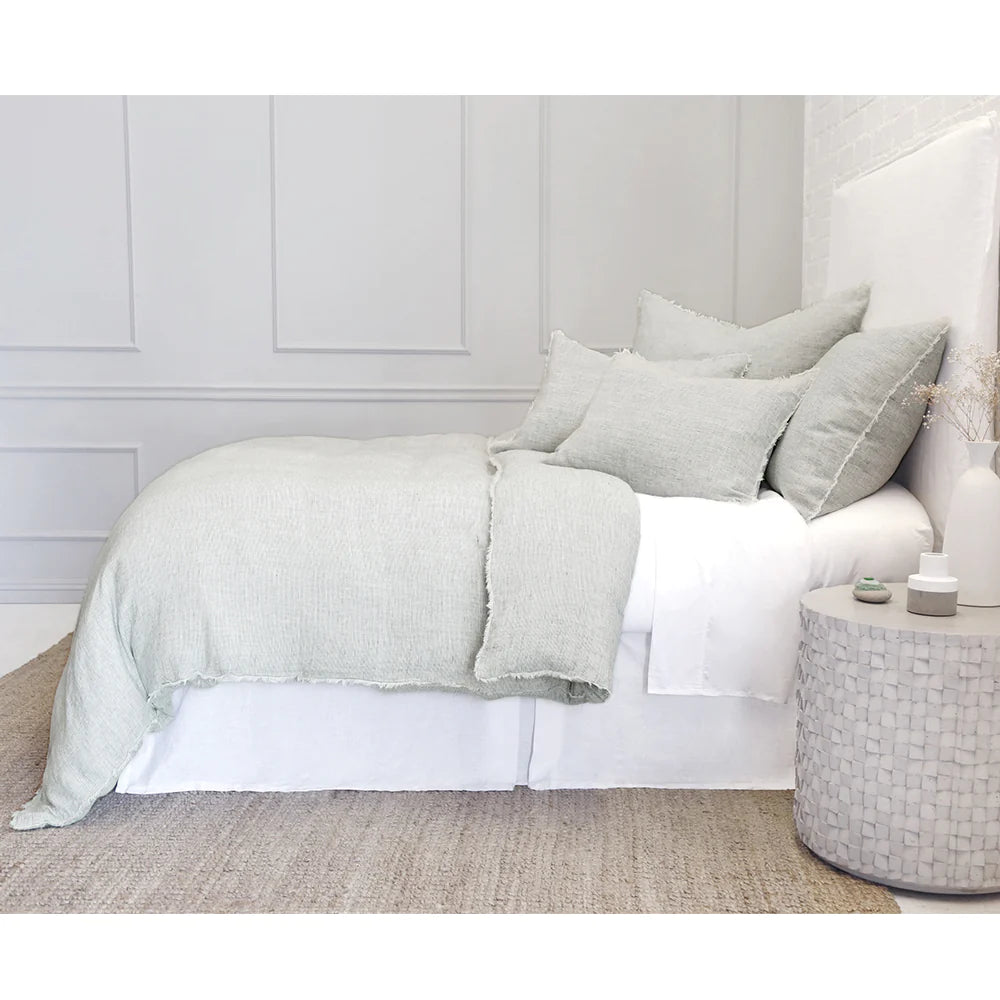 A neatly made bed adorned with sage green linens from the Logan Duvet set by POM POM stands against a light gray paneled wall. The bed is complemented by multiple pillows and a woven beige rug beneath. A textured white side table holds a small vase and decorative items, enhancing the room’s heathered effect.