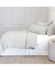 A neatly made bed adorned with sage green linens from the Logan Duvet set by POM POM stands against a light gray paneled wall. The bed is complemented by multiple pillows and a woven beige rug beneath. A textured white side table holds a small vase and decorative items, enhancing the room’s heathered effect.