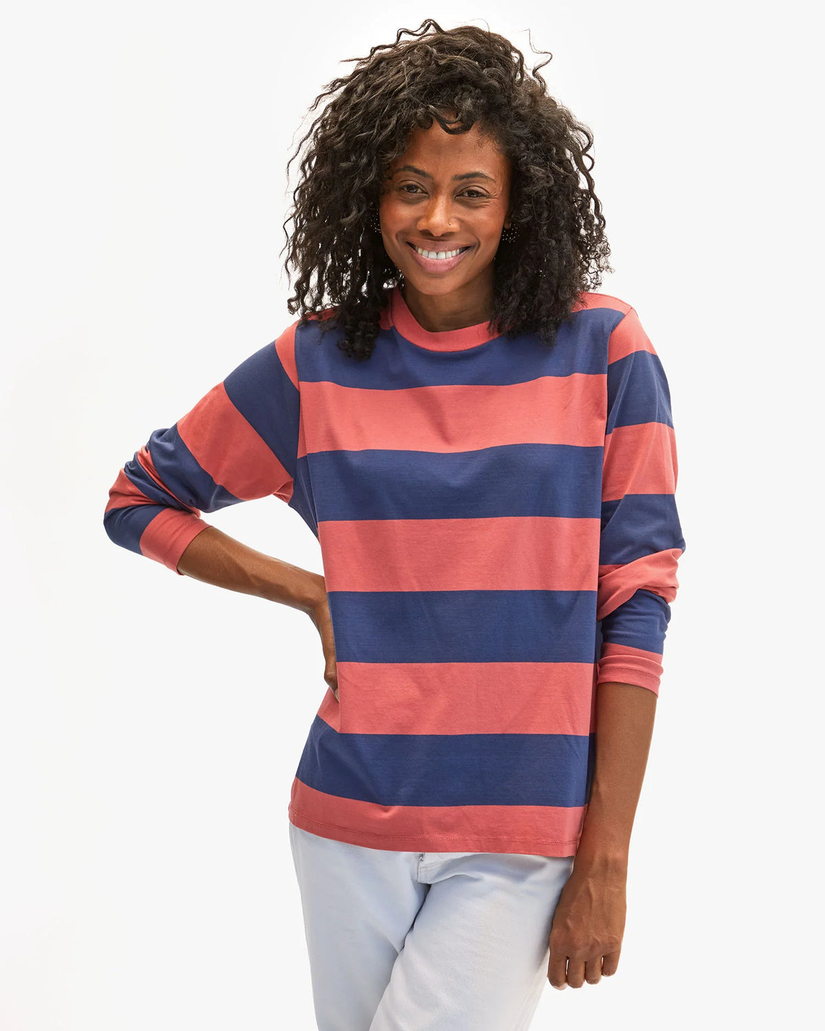 A person with curly hair smiles, wearing Clare Vivier's Long Sleeve Tee with wide red and navy blue horizontal stripes, inspired by classic Portuguese designs. They stand against a plain white backdrop with their right hand on their hip.