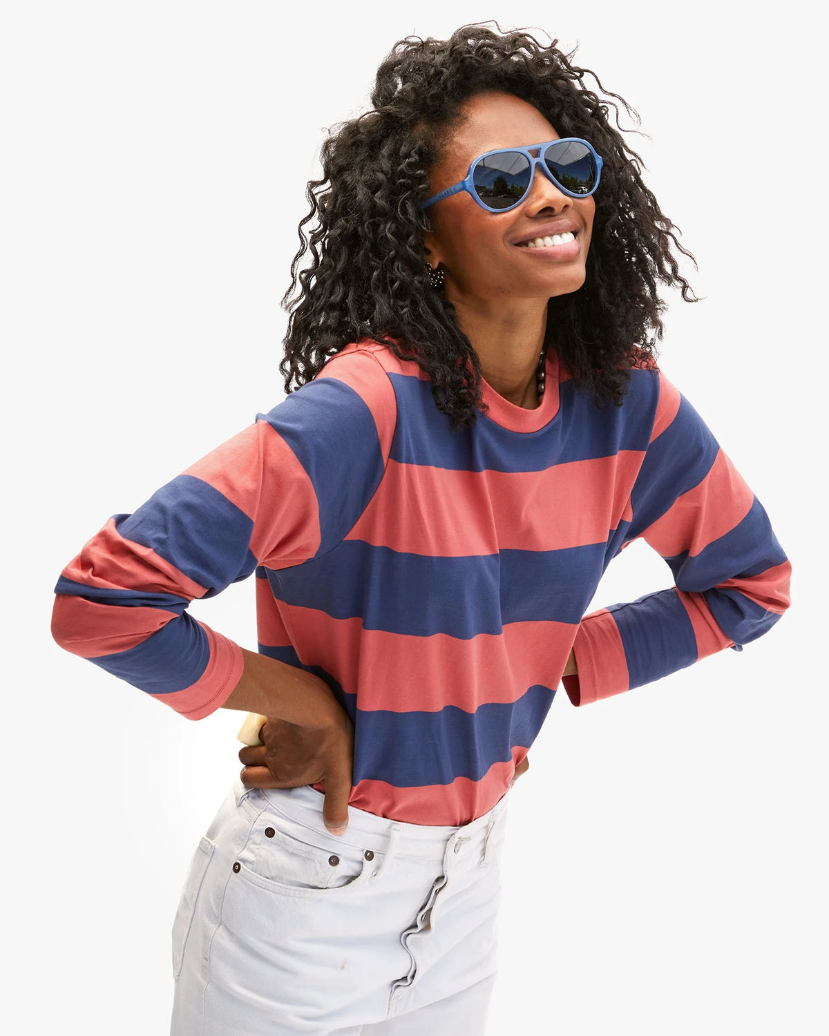 Wearing a Clare Vivier Long Sleeve Tee with blue and red stripes and blue sunglasses, a person smiles with hands on hips. Their curly hair frames their face as they stand confidently in white pants against a plain backdrop, exuding the casual charm of a sunny day in Portugal.