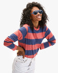 Wearing a Clare Vivier Long Sleeve Tee with blue and red stripes and blue sunglasses, a person smiles with hands on hips. Their curly hair frames their face as they stand confidently in white pants against a plain backdrop, exuding the casual charm of a sunny day in Portugal.