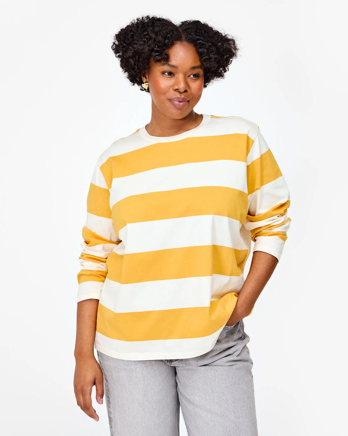 The individual is dressed in a yellow and white Long Sleeve Tee by Clare Vivier, made from 100% cotton, paired with gray pants. They stand against a plain white backdrop with a relaxed expression and hands in pockets.