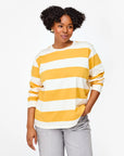The individual is dressed in a yellow and white Long Sleeve Tee by Clare Vivier, made from 100% cotton, paired with gray pants. They stand against a plain white backdrop with a relaxed expression and hands in pockets.