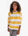 A curly-haired person smiles with closed eyes, wearing a Clare Vivier Long Sleeve Tee with wide yellow and white stripes described as reminiscent of sunny Portugal, paired with faded jeans against a plain white background.