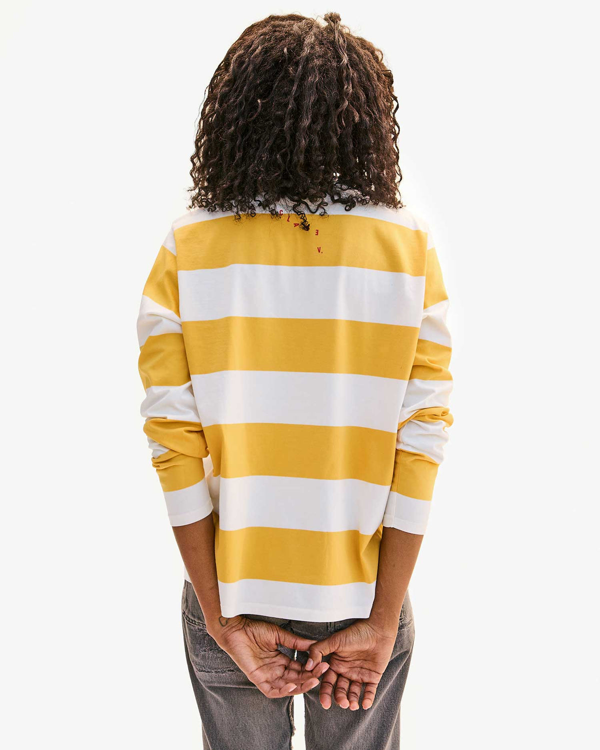 A person with curly hair, viewed from behind, wears a 100% cotton Clare Vivier yellow and white striped long sleeve tee and gray pants, holding their hands behind their back.