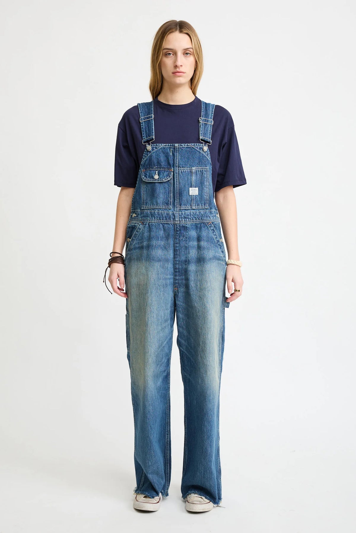 A person with long hair wears a navy tee and Denimist SLOUCH BIB OVERALL in Esher blue featuring a utility pocket, standing forward against a plain white background with a neutral expression and sneakers.