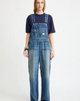 A person with long hair wears a navy tee and Denimist SLOUCH BIB OVERALL in Esher blue featuring a utility pocket, standing forward against a plain white background with a neutral expression and sneakers.
