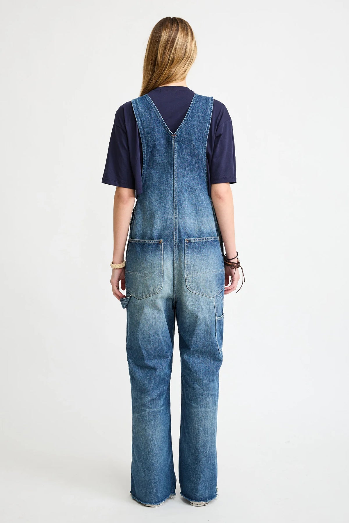 A person with long blonde hair faces away, wearing a dark T-shirt and Denimist’s SLOUCH BIB OVERALL in esher blue denim with back utility pockets. The background is plain white.