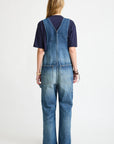 A person with long blonde hair faces away, wearing a dark T-shirt and Denimist’s SLOUCH BIB OVERALL in esher blue denim with back utility pockets. The background is plain white.