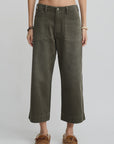 A person wearing Denimist’s Georgia Work Pant in olive green, featuring wide legs, 100% cotton fabric, and large front pockets, styled with a white cropped top and brown moccasin-style shoes against a plain light background.