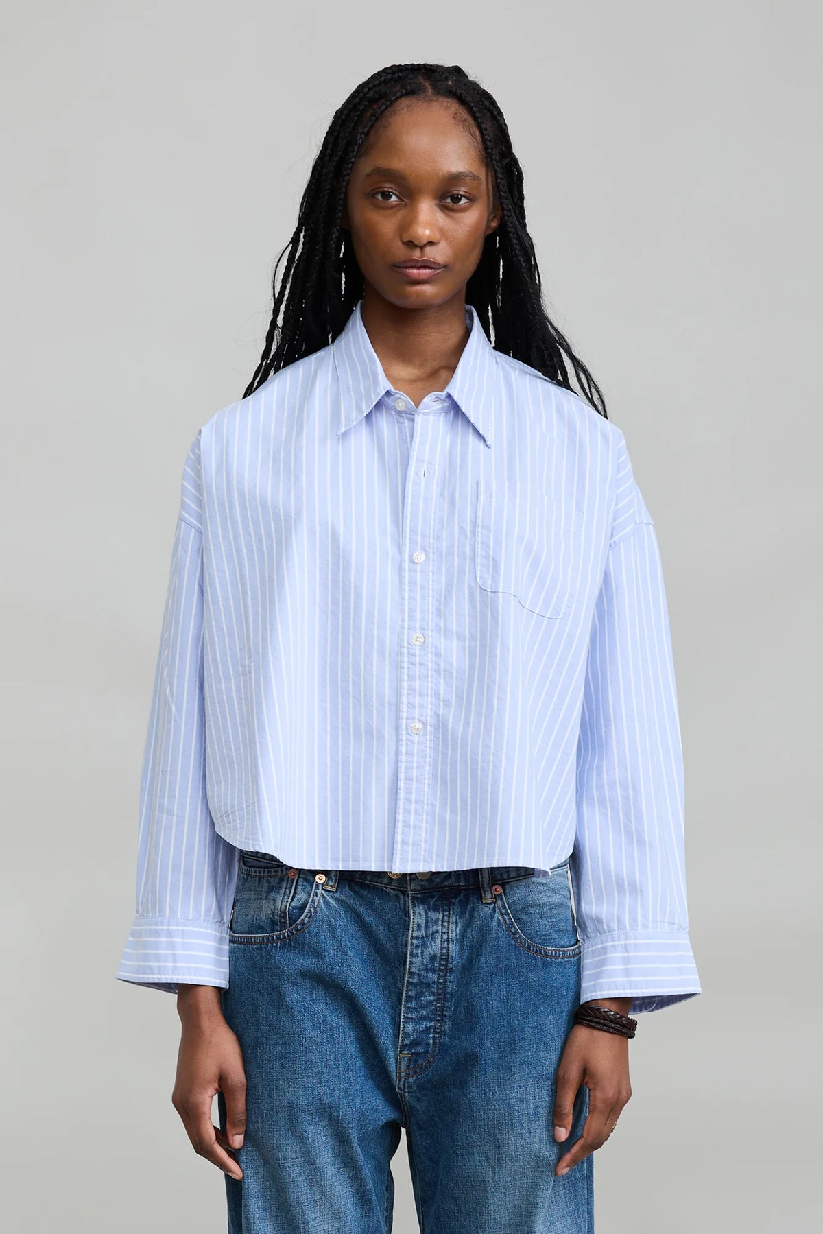 Denimist Bailey Cropped Stripe Oxford shirt in blue stripes styled with jeans, shown against a gray background.