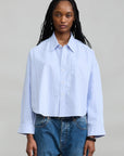 Denimist Bailey Cropped Stripe Oxford shirt in blue stripes styled with jeans, shown against a gray background.