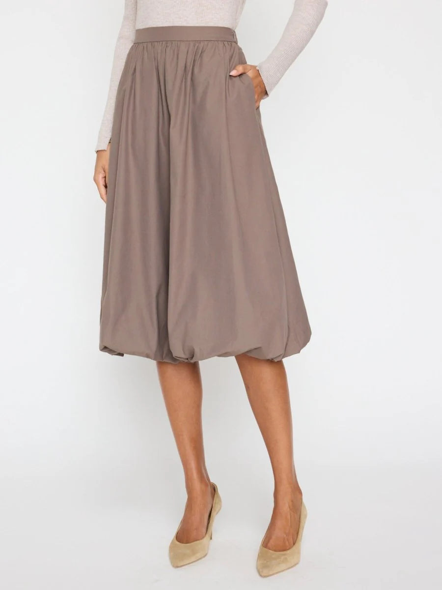 A person wears the BROCHU WALKER LOTI BUBBLE SKIRT in taupe, featuring a knee-length cut and elastic waistband, styled with a light long-sleeve top and beige low-heel shoes, standing against a white background for a modern look.