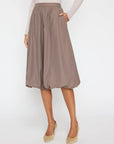 A person wears the BROCHU WALKER LOTI BUBBLE SKIRT in taupe, featuring a knee-length cut and elastic waistband, styled with a light long-sleeve top and beige low-heel shoes, standing against a white background for a modern look.