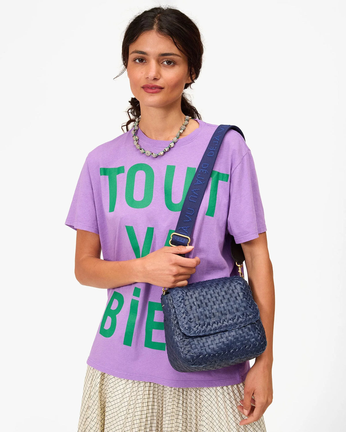 A woman in a lavender T-shirt, cream pleated skirt, and beaded necklace holds a blue bag with the Clare Vivier Adjustable Crossbody Strap, featuring cotton webbing, standing against a white background.