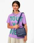 A woman in a lavender T-shirt, cream pleated skirt, and beaded necklace holds a blue bag with the Clare Vivier Adjustable Crossbody Strap, featuring cotton webbing, standing against a white background.