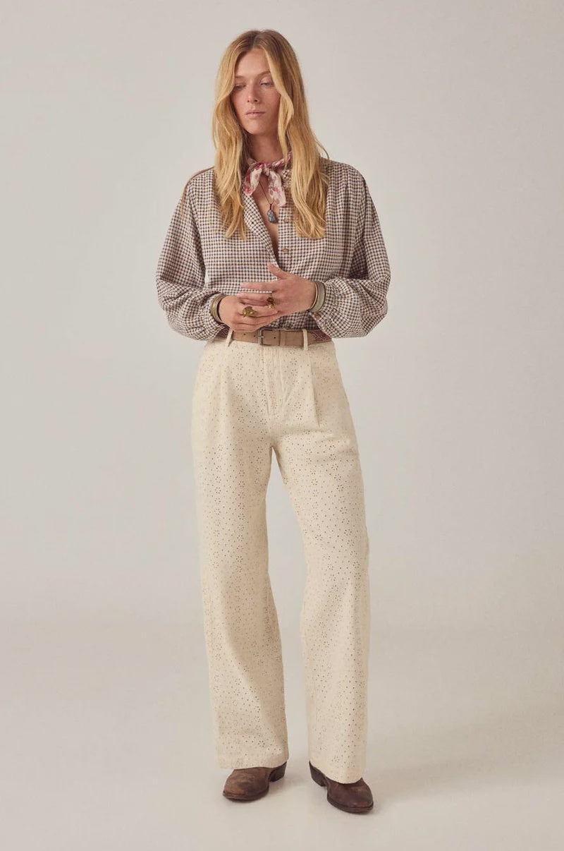 A woman stands against a plain background, wearing Maison Hotel's Theodore Pant in cream embroidered cotton twill with an eyelet pattern, a checked long-sleeve blouse, brown shoes, and a patterned neck scarf. Her hands are clasped and her blond hair is loose.