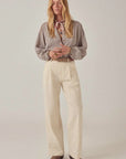 A woman stands against a plain background, wearing Maison Hotel's Theodore Pant in cream embroidered cotton twill with an eyelet pattern, a checked long-sleeve blouse, brown shoes, and a patterned neck scarf. Her hands are clasped and her blond hair is loose.