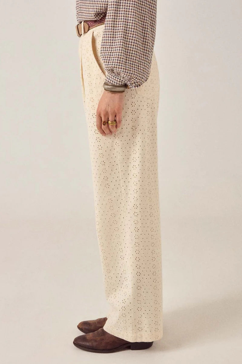 A person stands sideways against a plain light background, wearing Maison Hotel's cream-colored Theodore Pant—high-waisted, wide-leg with an eyelet pattern—paired with a checked long-sleeve shirt, brown leather shoes, a tan belt, and gold rings.