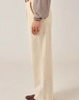 A person stands sideways against a plain light background, wearing Maison Hotel's cream-colored Theodore Pant—high-waisted, wide-leg with an eyelet pattern—paired with a checked long-sleeve shirt, brown leather shoes, a tan belt, and gold rings.