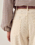 Seen from the waist down and behind, a person wears Maison Hotel's Theodore Pant—cream high-waisted wide-leg trousers in embroidered cotton twill with a floral eyelet pattern—paired with a checked shirt and burgundy patterned belt.