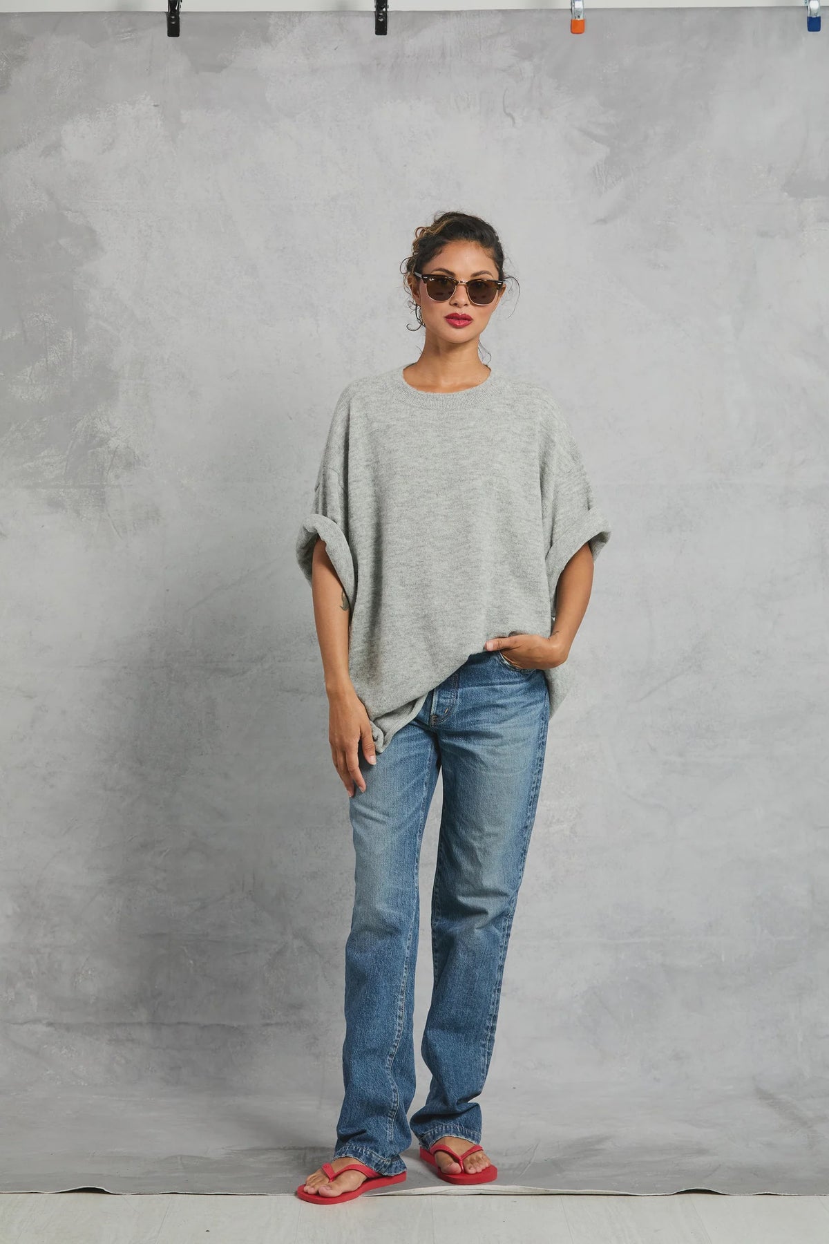 A woman poses before a gray textured backdrop, wearing sunglasses, blue jeans, red flip-flops, and the Mamene Boyfriend Tee—a loose, lightweight knit gray top. Her hair is tied up and one hand rests in her pocket.