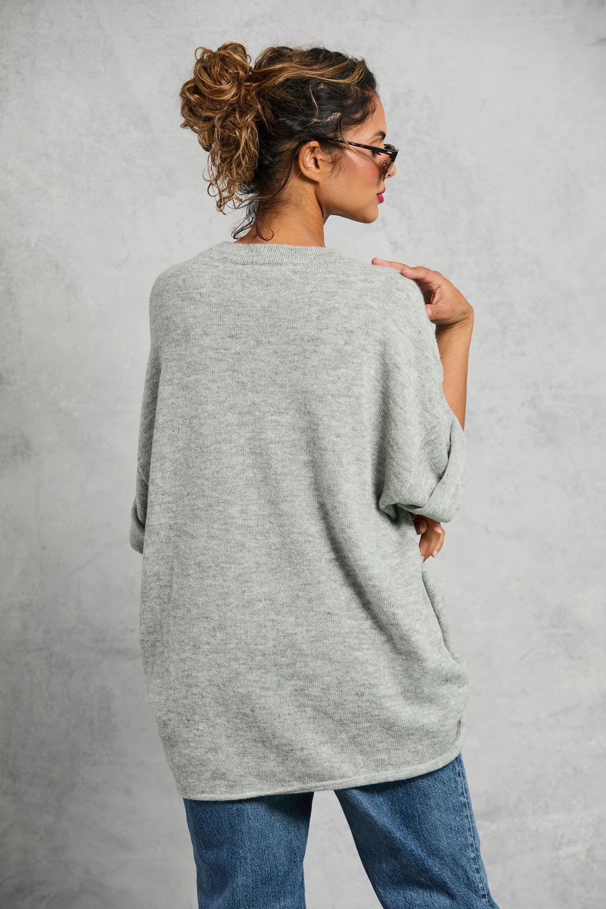 A woman with curly hair in a messy bun models the Mamene Boyfriend Tee, paired with blue jeans and glasses, standing with her back to the camera and turning her head to the side against a light gray background.
