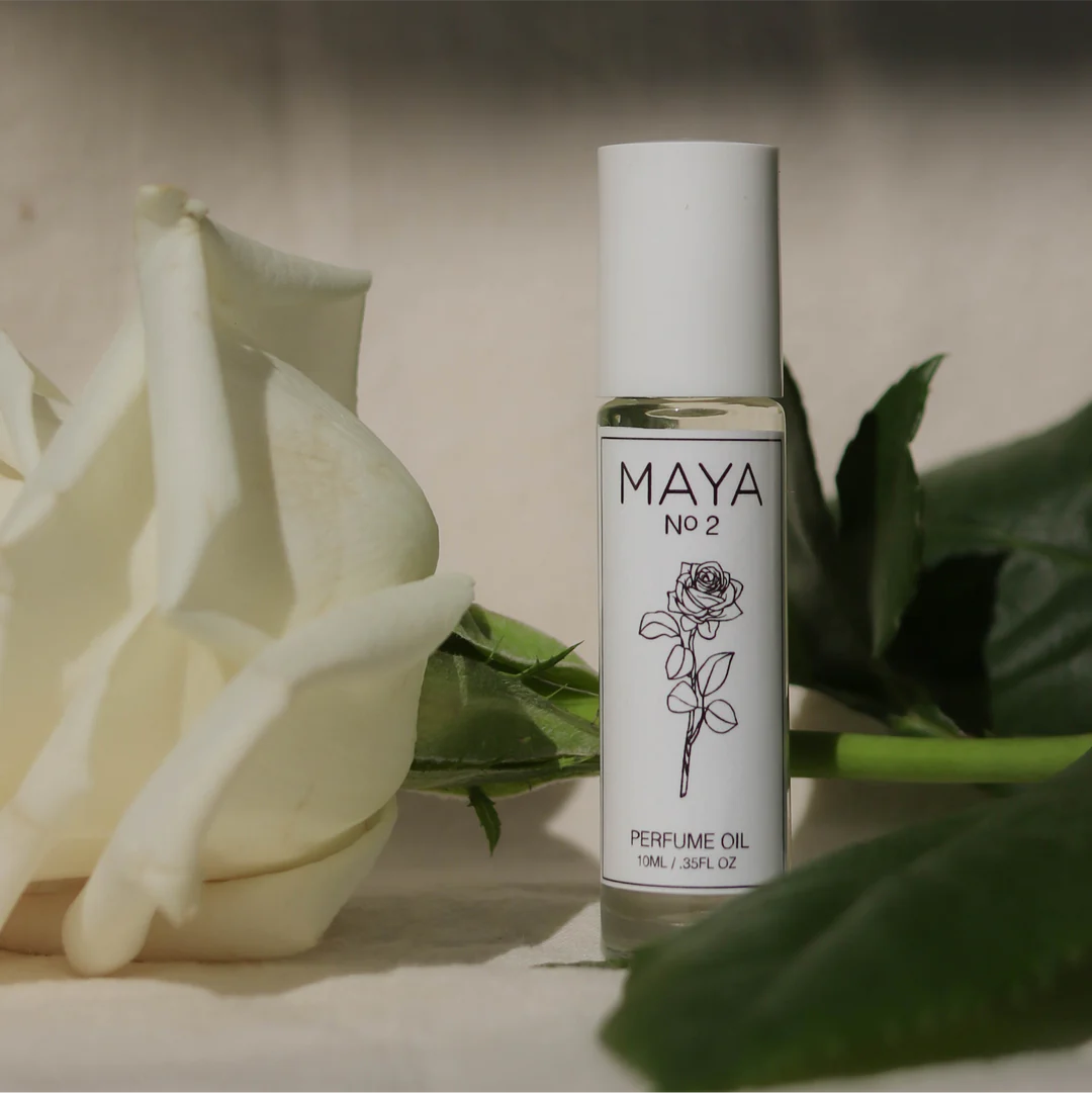 A white MAYA NO.2 10ML bottle by Maya Fragrances, featuring a rose illustration, stands upright beside a white rose and green leaves on a neutral background, evoking the sexy, musky undertones of this perfume oil.