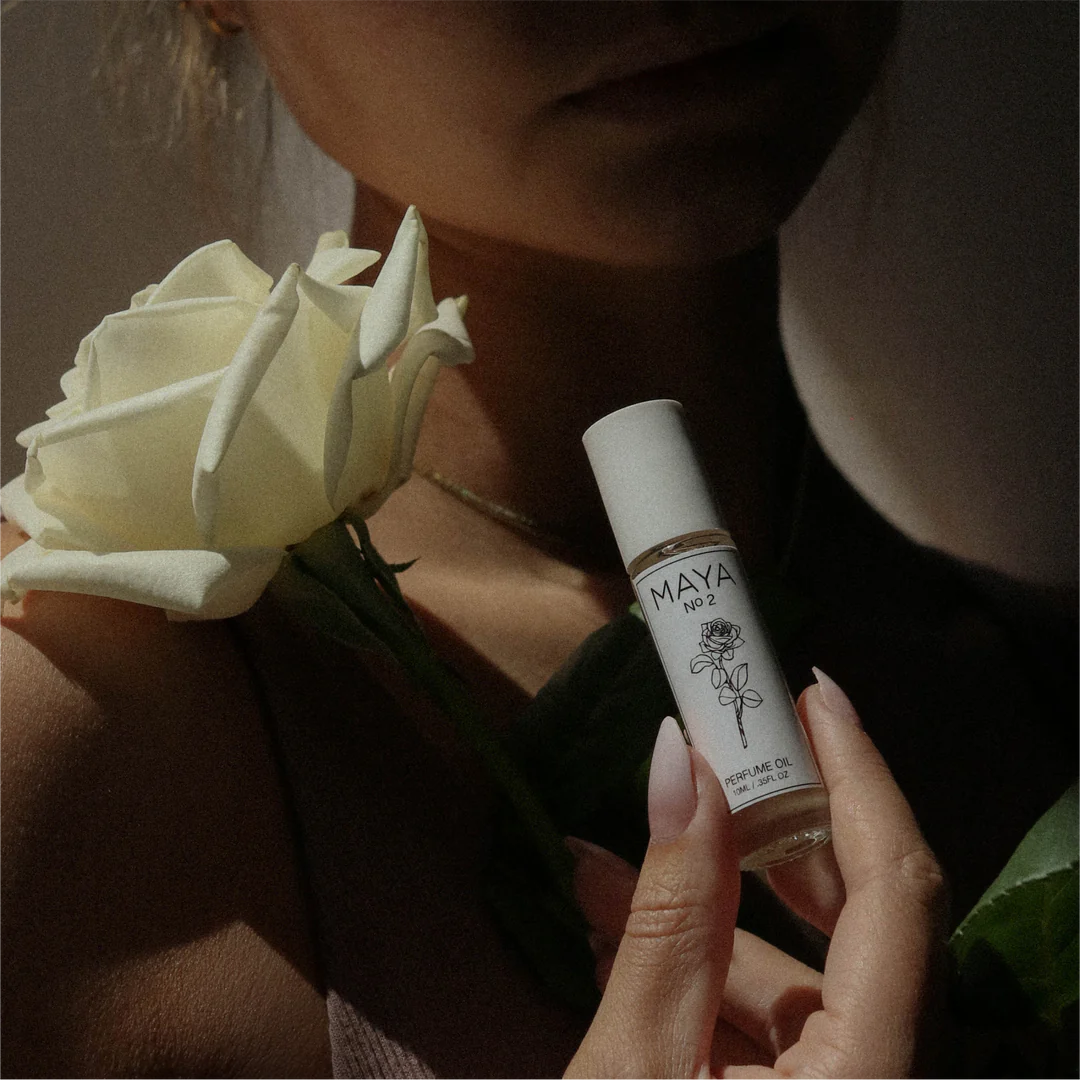 A person holds a white rose and a 10ML bottle of Maya Fragrances’ MAYA NO.2 perfume oil, its minimalist floral design and white cap catching soft natural light as the musky scent unveils itself in gentle shadows.