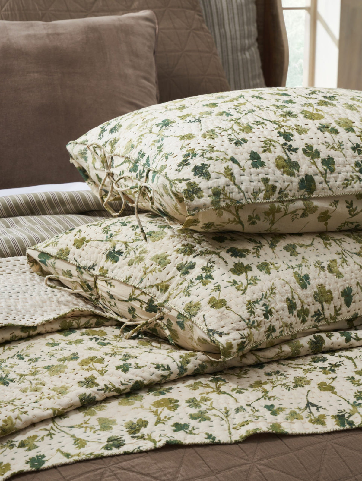 Two Amity Home Millie Euro Shams with green and beige floral pillowcases are stacked on a hand-quilted cotton voile quilt, placed over brown and striped bedding in a softly lit room.