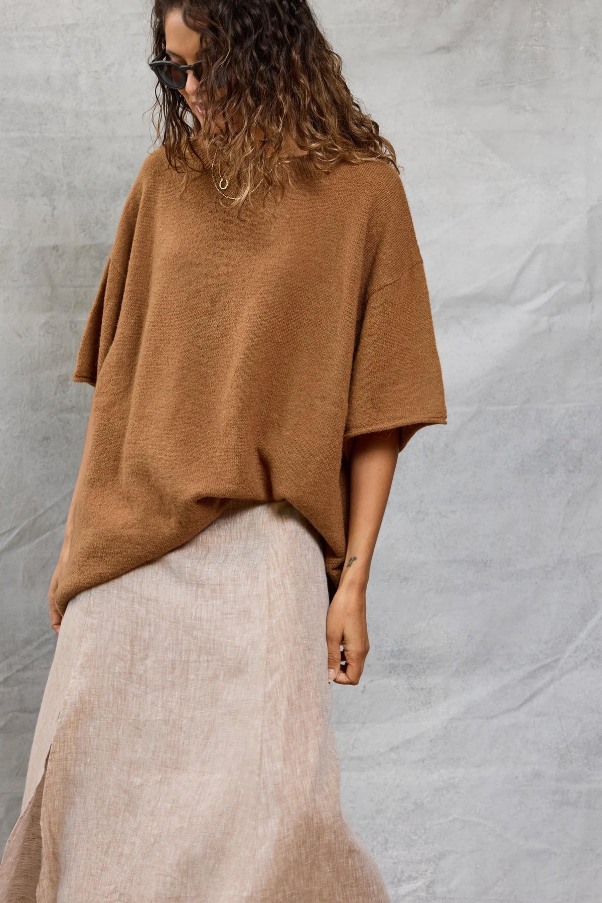 A person wearing Mamene's Boyfriend Tee, a loose brown lightweight knit short-sleeve sweater, with a long beige skirt stands against a light gray textured background, wavy hair down and one hand by their side.