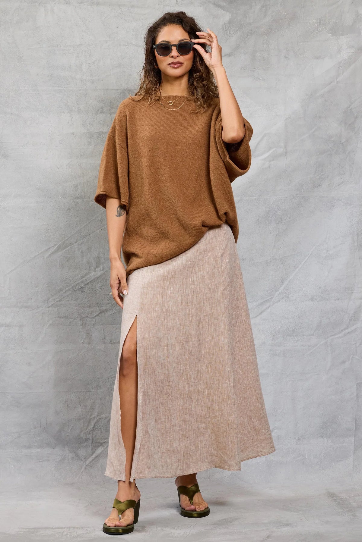 A woman in wavy hair and sunglasses poses against a textured gray background, wearing the Mamene Boyfriend Tee, a loose brown lightweight knit top, paired with a light beige thigh-high slit maxi skirt and green platform sandals.