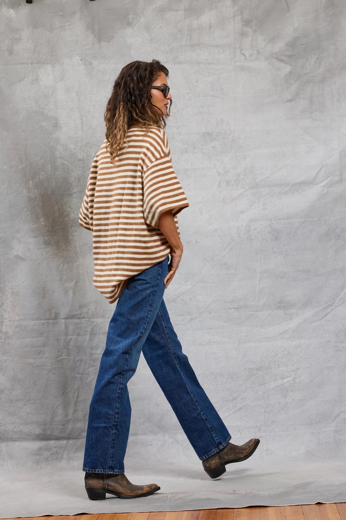 Wearing Mamene's Boyfriend Tee, a person with wavy hair, sunglasses, blue jeans, and brown boots walks before a textured gray backdrop.