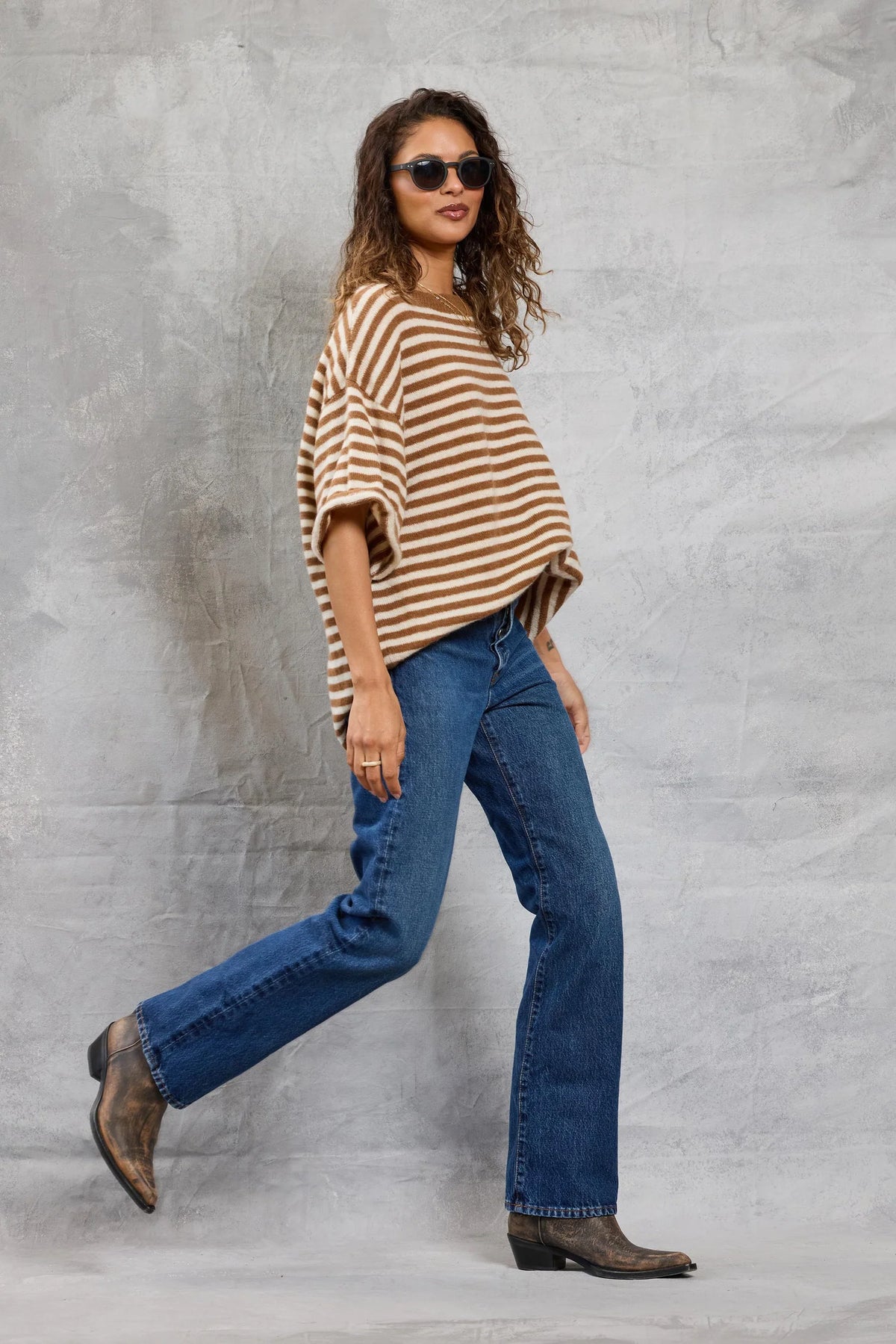 A woman wearing sunglasses, blue jeans, brown boots, and Mamene's Boyfriend Tee walks confidently in front of a textured gray background.