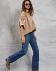 A woman wearing sunglasses, blue jeans, brown boots, and Mamene's Boyfriend Tee walks confidently in front of a textured gray background.