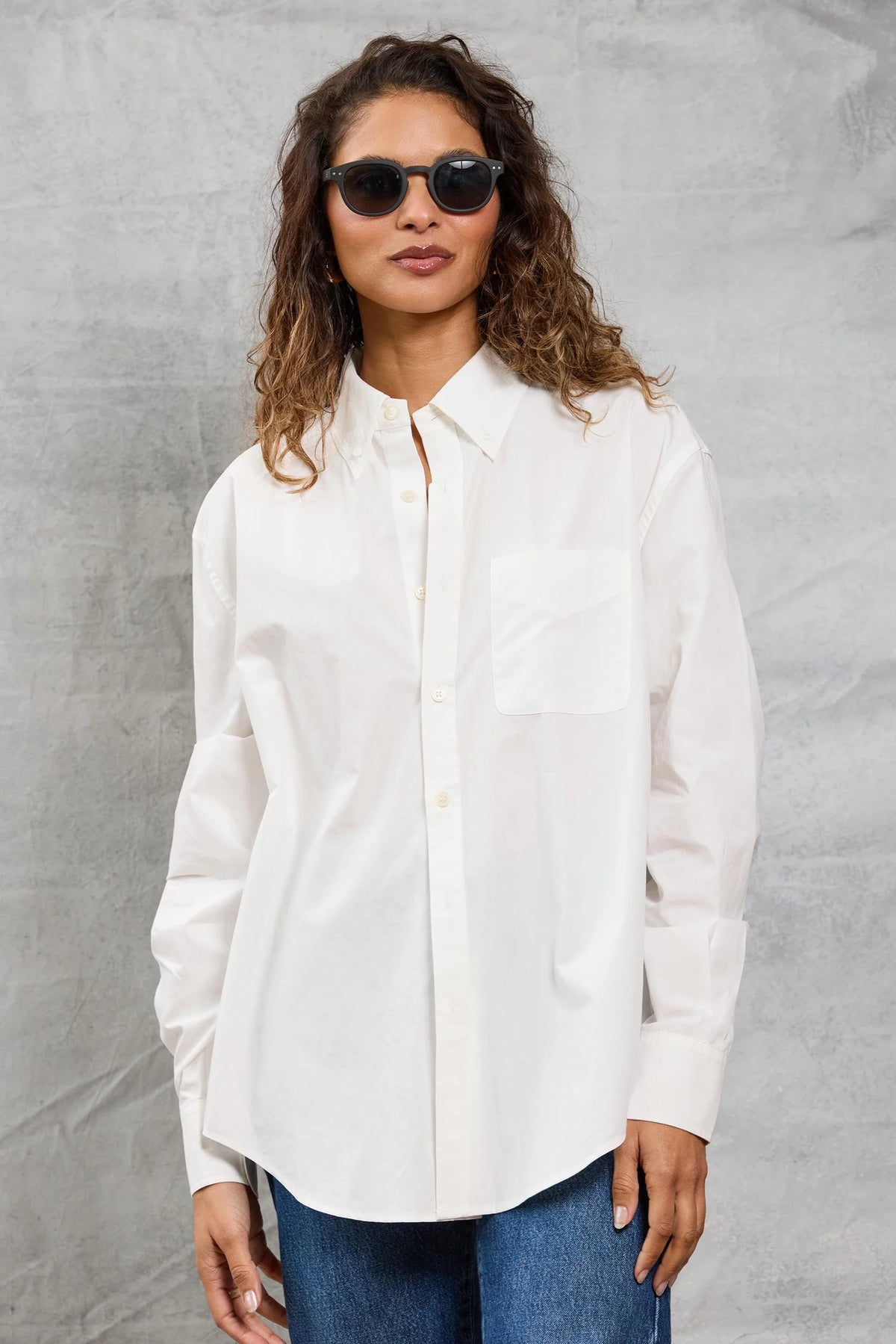 A woman with wavy brown hair and black sunglasses wears the Mamene Slim Repeat Shirt and blue jeans, standing against a light gray textured background.