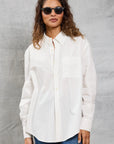 A woman with wavy brown hair and black sunglasses wears the Mamene Slim Repeat Shirt and blue jeans, standing against a light gray textured background.