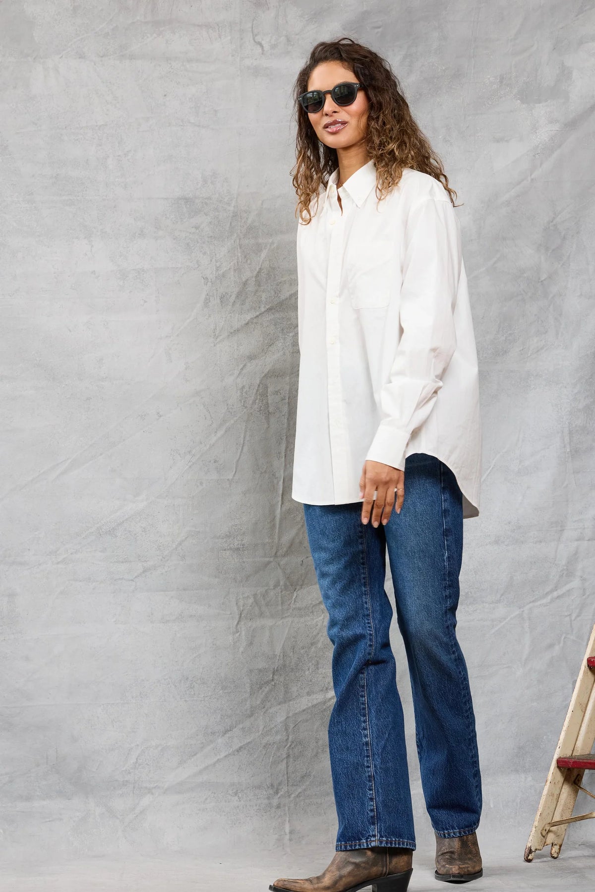 A person with long curly hair, sunglasses, blue jeans, and brown boots models the Mamene Slim Repeat Shirt against a light gray textured background near a wooden ladder.