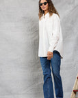 A person with long curly hair, sunglasses, blue jeans, and brown boots models the Mamene Slim Repeat Shirt against a light gray textured background near a wooden ladder.