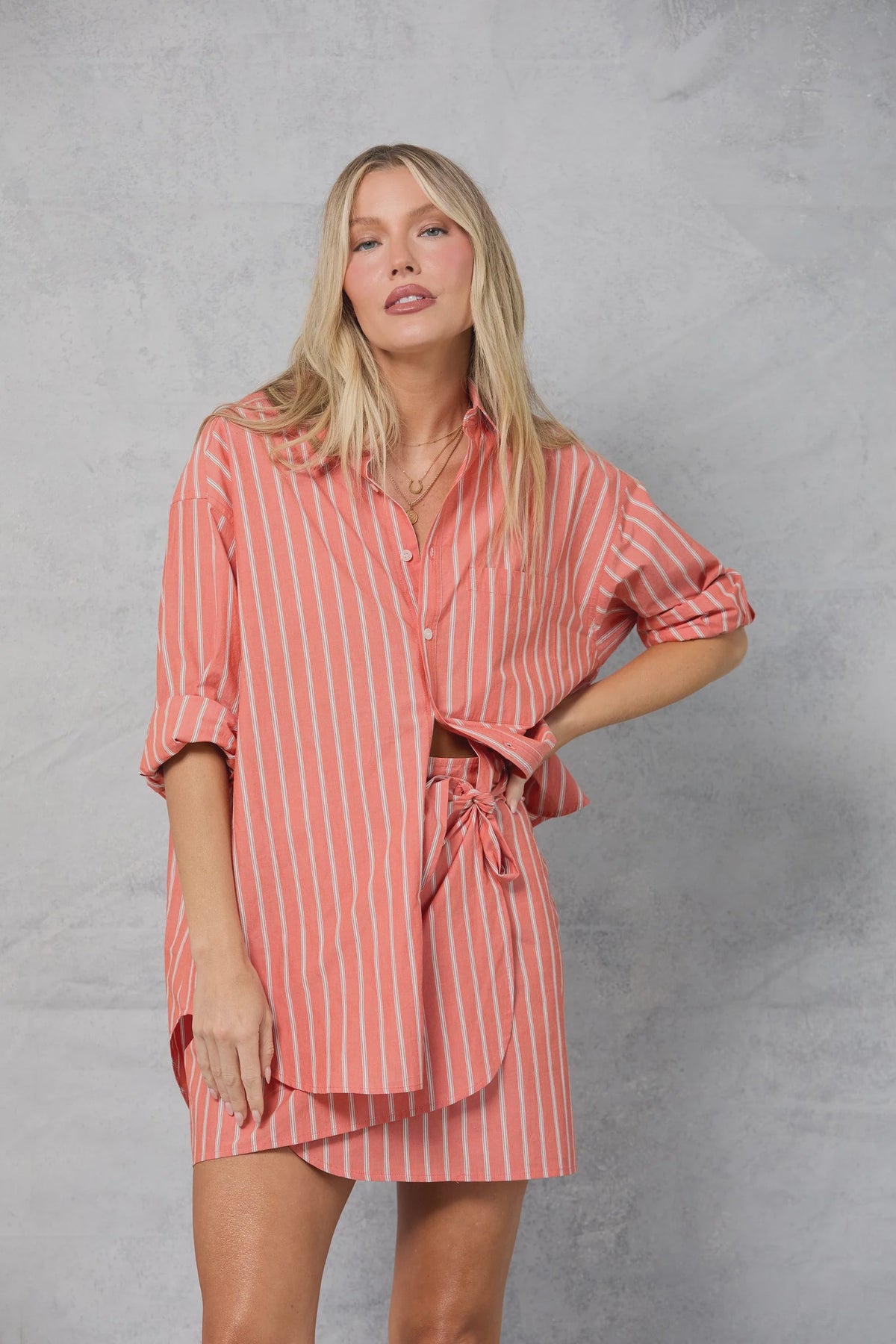 A woman with long blonde hair wears the Mamene Repeat Shirt—a loose-fitting, pink and white Japanese yarn-dye stripe cotton button-down—paired with matching shorts, standing against a light gray textured background, one hand on her hip.