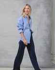 A blonde woman walks confidently in the Mamene Slim Repeat Shirt and dark navy wide-leg pants with open-toe sandals, set against a neutral mottled grey studio backdrop.
