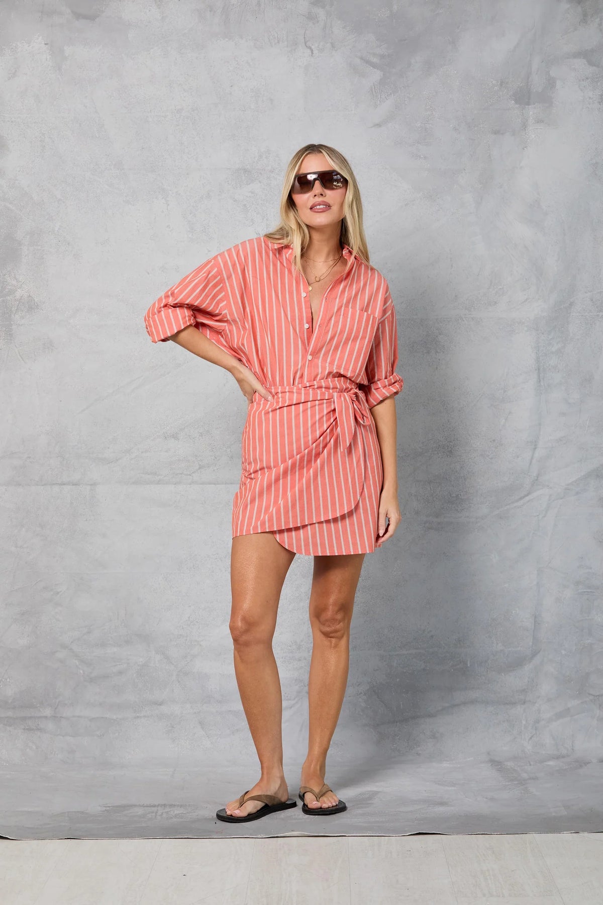 A woman in sunglasses, a coral and white striped shirt dress tied at the waist, black flip-flops, and the Mamene Repeat Wrap Tennis Skirt stands before a gray textured backdrop, creating a playful warm-weather vibe.
